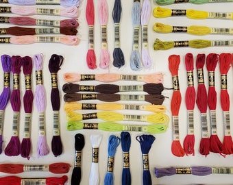 May include: A collection of embroidery floss spools in various colors, including red, blue, green, yellow, purple, and white. The spools are arranged in rows and columns on a white surface.