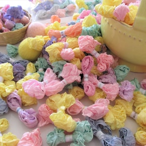 May include: A collection of pastel-colored fabric scraps tied into bows. The fabric scraps are in shades of pink, yellow, blue, and green. The fabric scraps are arranged in a pile on a white surface.