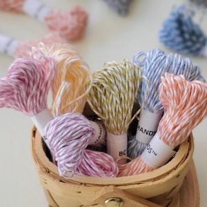 May include: A small, woven basket filled with colorful embroidery floss. The floss is in shades of pink, yellow, green, and blue. Some spools have the text "HANDS" and "BC" on the labels. Other spools are scattered in the background.