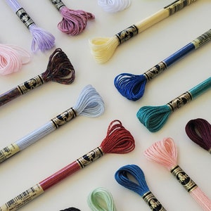 May include: Assortment of DMC embroidery floss spools in various colours, including pink, lavender, white, yellow, green, blue, red, and black. Each spool has a gold label with the DMC logo and the thread number.