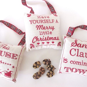 May include: Three white fabric ornaments with red embroidered text. The ornaments have red and black plaid ribbon loops. The text on the ornaments reads "This House Believes", "Have Yourself A Merry Little Christmas", and "Santa Claus Is Coming To Town".