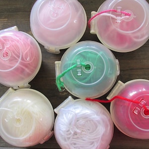 May include: A selection of yarn in transparent plastic containers. The yarn is in shades of pink, white, and green. The containers are round with a small opening for the yarn to come out.