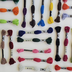 May include: Assortment of embroidery floss spools in various colors, including red, green, blue, purple, brown, and yellow. The spools are arranged in a grid pattern on a white surface.