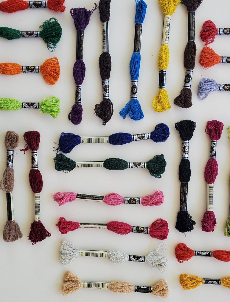 Puede incluir: Surtido de carretes de hilo de bordar en varios colores, incluyendo rojo, verde, azul, morado, marr&oacute;n y amarillo. Los carretes est&aacute;n dispuestos en un patr&oacute;n de cuadr&iacute;cula sobre una superficie blanca.