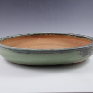May include: A shallow, round bonsai pot with a pale green exterior and a natural terracotta interior. The rim has a darker, weathered appearance. The pot is designed for planting small trees and shrubs.