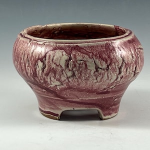 May include: A small, round ceramic pot with a maroon and cream marbled glaze. The pot has a wide opening, a rounded body, and three small feet. The interior is a darker maroon colour. This decorative pot is suitable for small plants.