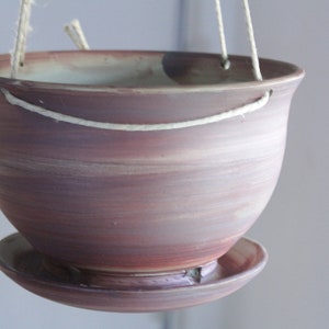 May include: A hanging planter made of brown and white glazed ceramic with a saucer. The planter has a rope hanging system.