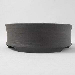 May include: A dark gray ceramic bonsai pot with a rounded shape and a slightly flared rim. The pot has three small feet on the bottom.