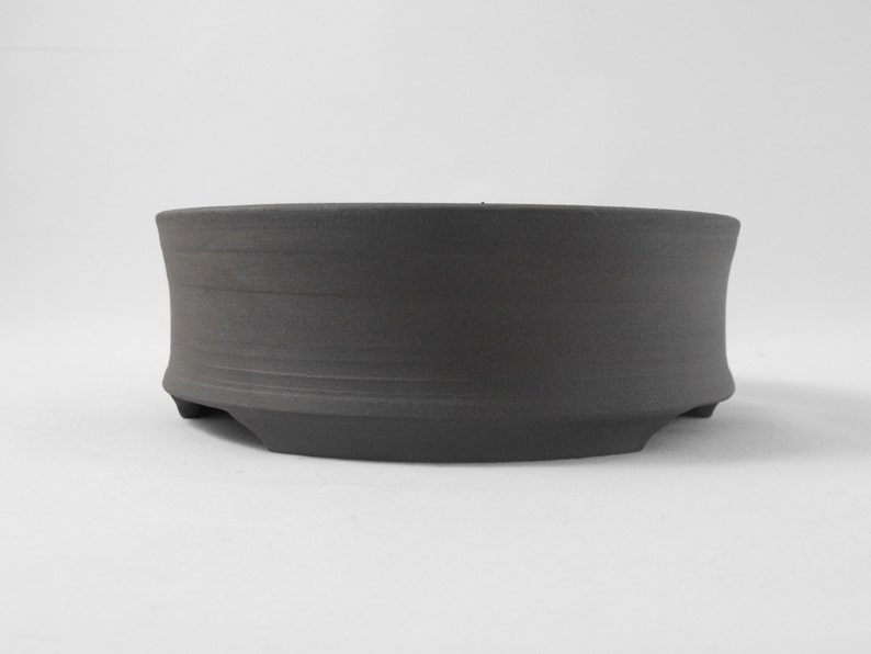 May include: A dark gray ceramic bonsai pot with a rounded shape and a slightly flared rim. The pot has three small feet on the bottom.