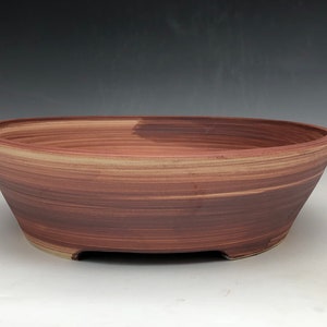 May include: A large, oval-shaped ceramic bonsai pot. The pot has a reddish-brown color with lighter brown horizontal brushstrokes. The pot has a wide, shallow design with a slightly flared rim and small feet.