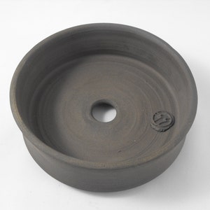 May include: A dark gray ceramic planter with a circular opening in the center. The planter has a smooth, slightly textured surface and a small, circular mark on the side.