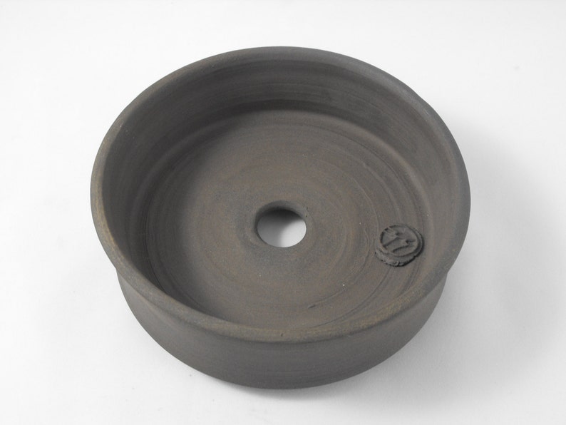 May include: A dark gray ceramic planter with a circular opening in the center. The planter has a smooth, slightly textured surface and a small, circular mark on the side.