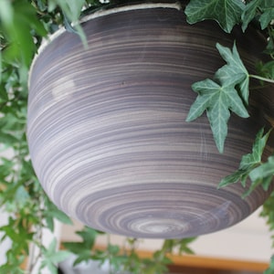 May include: A large, round, dark grey planter with a swirling pattern. The planter is hanging from a vine of green leaves.