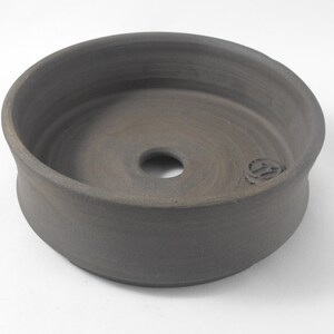 May include: A dark gray ceramic planter with a round, wide base and a slightly flared rim. The planter has a drainage hole in the center of the bottom.