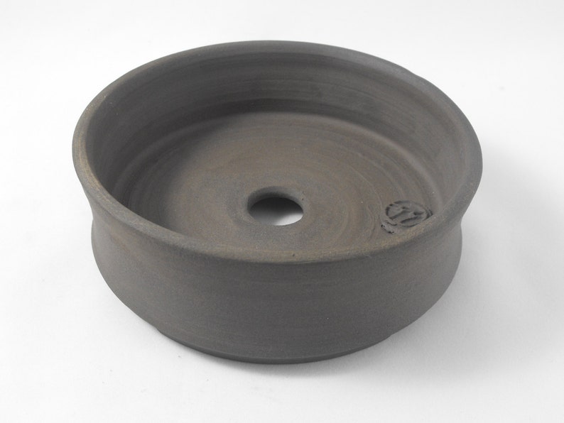 May include: A dark gray ceramic planter with a round, wide base and a slightly flared rim. The planter has a drainage hole in the center of the bottom.