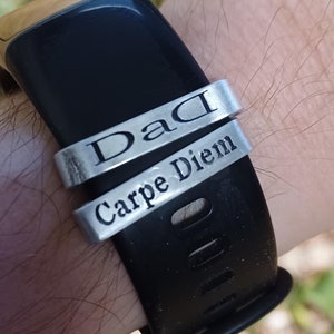 Könnte beinhalten: Ein schwarzes Smartwatch-Armband mit zwei silbernen Ringen. Der obere Ring trägt die Aufschrift "Dad" und der untere Ring "Carpe Diem" in einer einfachen Schriftart. Der Hintergrund ist verschwommen mit Grün und Gelb.