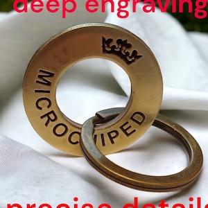 Op de afbeelding: Een gouden metalen sleutelhanger met een ronde ring en een grotere ring met de woorden "MICROCHIPPED" gegraveerd rond de rand. Een kroon is gegraveerd op de grotere ring.