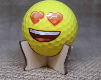 May include: A yellow golf ball with a smiley face and heart eyes. The golf ball is on a wooden stand.