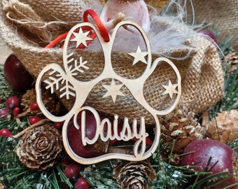 May include: Wooden paw print ornament with the name "Daisy" engraved on it. The ornament is decorated with snowflakes and stars. It is hanging from a red ribbon and is surrounded by a burlap bag and a small angel figurine.
