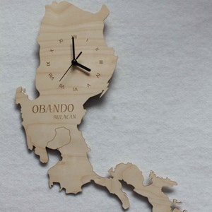 Può includere: Un orologio in legno a forma di contorno di Obando, Bulacan, Filippine. L'orologio ha lancette nere e numeri romani.