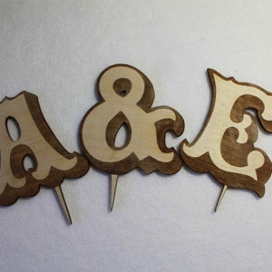May include: Three wooden cake toppers, each with a different letter. The letters are A, & and E. The letters are in a decorative font and are brown with a lighter wood color in the center.