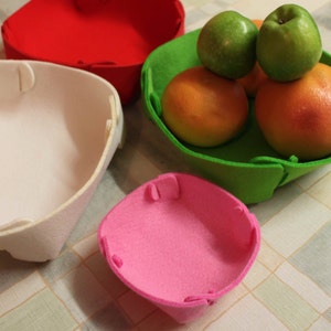 May include: Four felt bowls in different colors: white, red, green, and pink. The green bowl contains three green apples and two orange fruits.