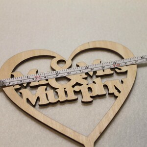 Puede incluir: Un adorno para pastel de madera en forma de coraz&oacute;n con el texto "Mr & Mrs Murphy" en una fuente decorativa. El adorno se est&aacute; midiendo con una cinta m&eacute;trica, lo que indica que tiene aproximadamente 10 cm de ancho.