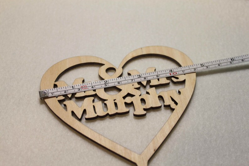 Puede incluir: Un adorno para pastel de madera en forma de coraz&oacute;n con el texto "Mr & Mrs Murphy" en una fuente decorativa. El adorno se est&aacute; midiendo con una cinta m&eacute;trica, lo que indica que tiene aproximadamente 10 cm de ancho.