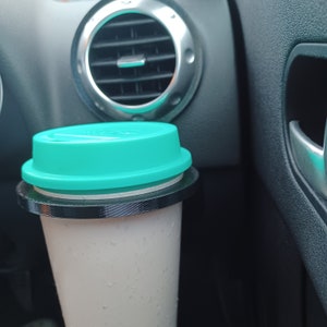 Op de afbeelding: Een witte reistumbler met een turquoise deksel en een zwarte ring rond de bovenkant. De tumbler staat in een bekerhouder in de auto.