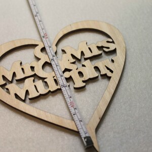 Puede incluir: Un adorno para pastel de madera en forma de coraz&oacute;n con las palabras "Mr & Mrs Murphy" recortadas. El adorno se est&aacute; midiendo con una cinta m&eacute;trica.