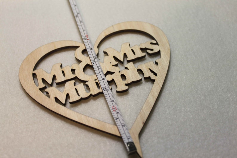 Puede incluir: Un adorno para pastel de madera en forma de coraz&oacute;n con las palabras "Mr & Mrs Murphy" recortadas. El adorno se est&aacute; midiendo con una cinta m&eacute;trica.