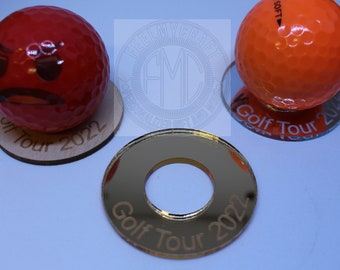 May include: Three golf ball display stands. Two are made of wood with the text "Golf Tour 2022" engraved on them. One is a gold mirror acrylic stand with the text "Golf Tour 2022" engraved on it.