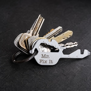May include: A silver multi-tool key with the words "Mr. Fix It" engraved on it, attached to a keyring with several keys. The multi-tool has various functions, including a ruler, and is set against a dark background.