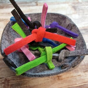 Pode incluir: Uma tigela de feltro cinza cheia de tiras de feltro coloridas. As tiras são de várias cores, incluindo vermelho, verde, rosa, azul e preto. As tiras são dispostas em um padrão aleatório, criando um design visualmente atraente e texturizado.