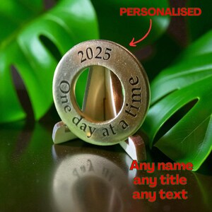 Può includere: Un gettone circolare color oro con l'anno 2025 inciso in alto. Le parole "One day at a time" sono incise attorno al bordo. Il gettone è esposto su un piccolo supporto di legno, con il testo "Any name any title any text" visibile.