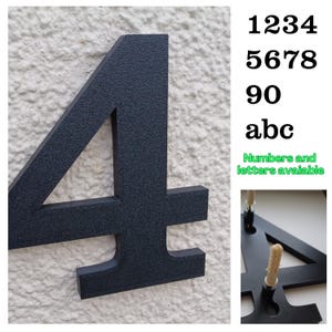 Può includere: Un numero civico 4 in metallo nero montato su un muro bianco strutturato. Il numero è grande e in grassetto, con una texture leggermente ruvida. Sono disponibili numeri e lettere aggiuntivi.