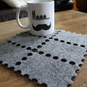 May include: White ceramic mug with a black mustache and the word "Mmmm..." printed on it. The mug sits on a gray felt placemat with a scalloped edge and dotted cutouts. The mug and placemat are on a wooden table.