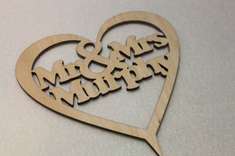 Puede incluir: Un adorno para pastel de madera en forma de coraz&oacute;n con el texto "Mr & Mrs Murphy" recortado.