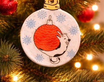 Cat hanging from ornament while it snows ornament