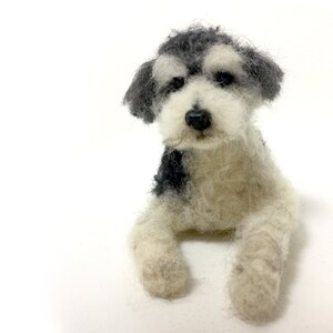 May include: A handmade felt dog figurine in a seated position. The dog has a black nose, black and gray fur on its head and back, and white fur on its body and legs. The dog is set against a white background.
