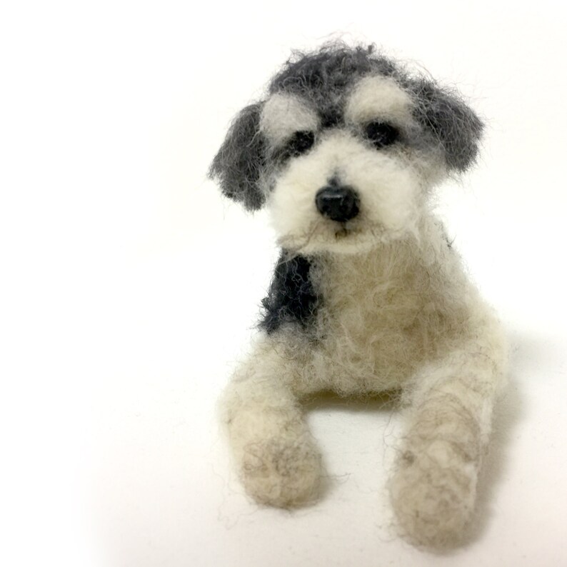 May include: A handmade felt dog figurine in a seated position. The dog has a black nose, black and gray fur on its head and back, and white fur on its body and legs. The dog is set against a white background.