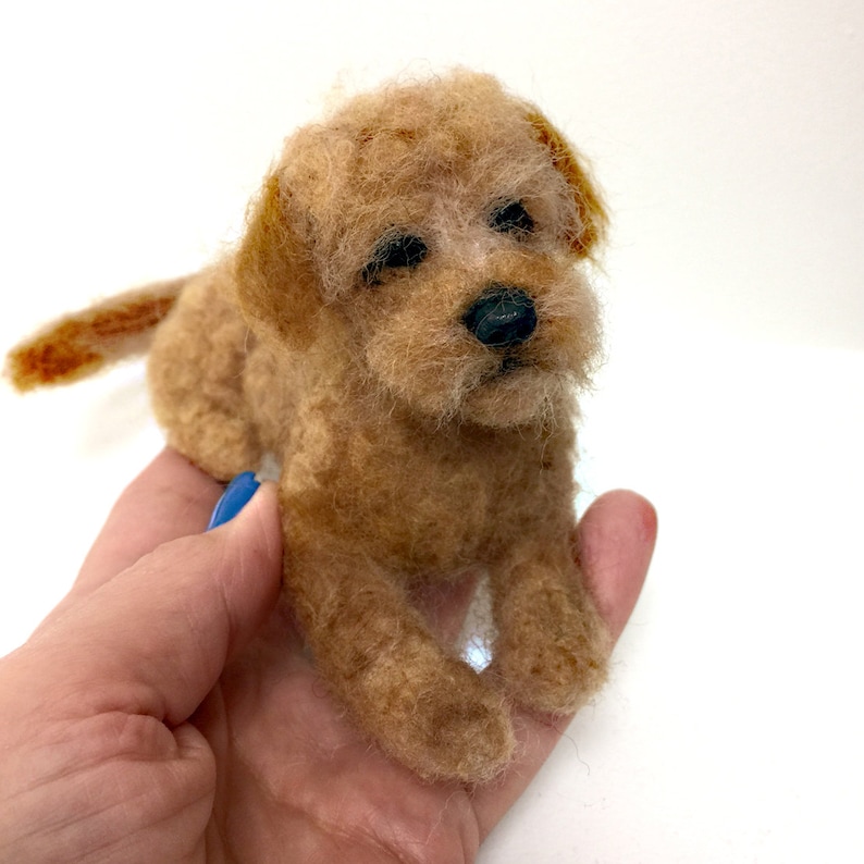 May include: A handmade, tan-colored felted dog figurine. The small dog has a fluffy texture, black eyes, and a black nose. The dog is lying down with its tail extended. The figurine is held in a hand against a white background.