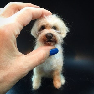May include: A small, handmade, off-white dog figurine with a brown nose and eyes. The dog is made of wool and has a fluffy appearance. The figurine is held by a hand with a blue painted nail.