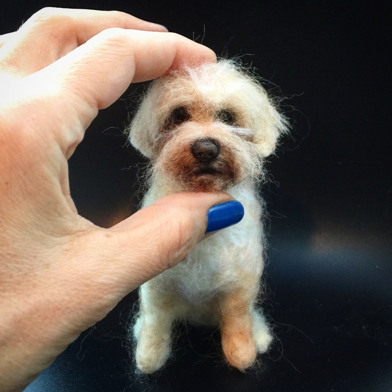 May include: A small, handmade, off-white dog figurine with a brown nose and eyes. The dog is made of wool and has a fluffy appearance. The figurine is held by a hand with a blue painted nail.