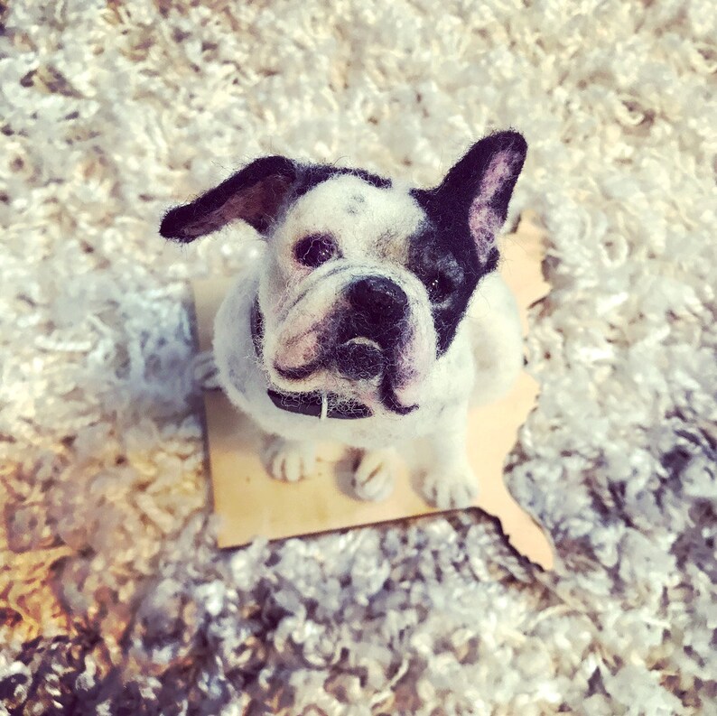 May include: A white and black French bulldog figurine made of felt. The figurine is sitting on a wooden base and is looking up at the camera.