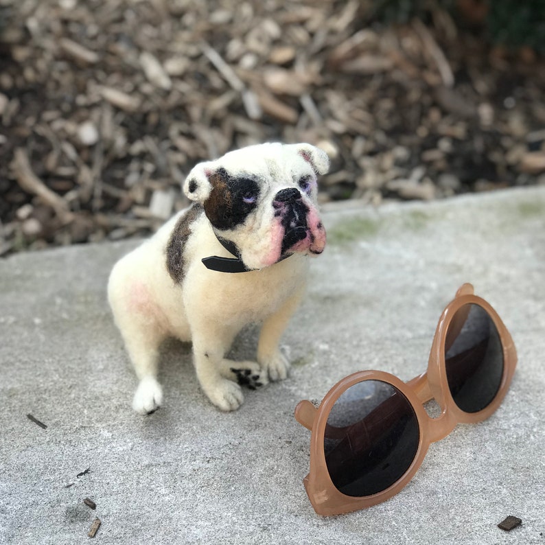 May include: A white and brown felt bulldog figurine with black eyes and a black collar. The figurine is sitting on a gray surface next to a pair of brown sunglasses.