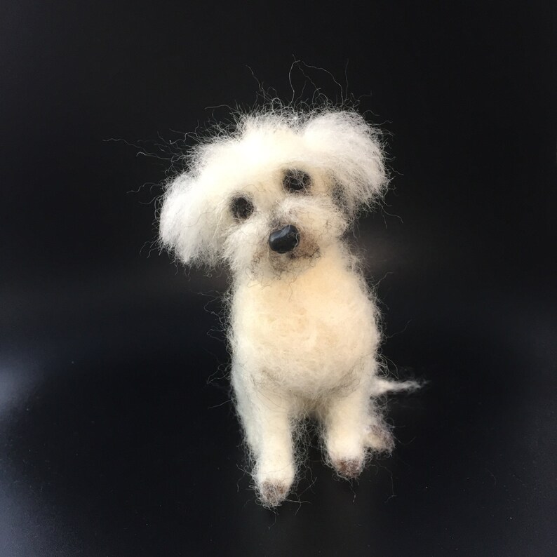 May include: A handmade, off-white felted dog figurine with a fluffy coat and a black nose. The dog is sitting, with its head tilted slightly. The background is a solid black.