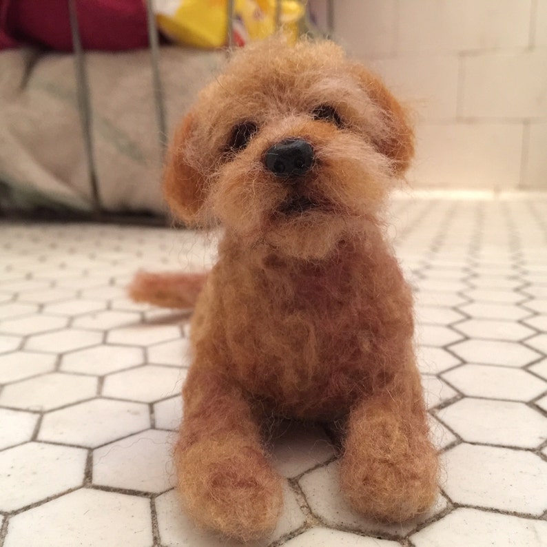 May include: A handcrafted, miniature, brown felted dog figurine. The dog has a fluffy, textured appearance with a black nose and small, dark eyes. It is posed sitting on a white tile floor with a hexagonal pattern.