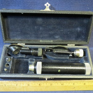 May include: A vintage ophthalmoscope in a black leather case with a wooden ruler for scale. The ophthalmoscope has a black metal body and a silver lens.