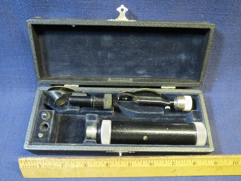 May include: A vintage ophthalmoscope in a black leather case with a wooden ruler for scale. The ophthalmoscope has a black metal body and a silver lens.